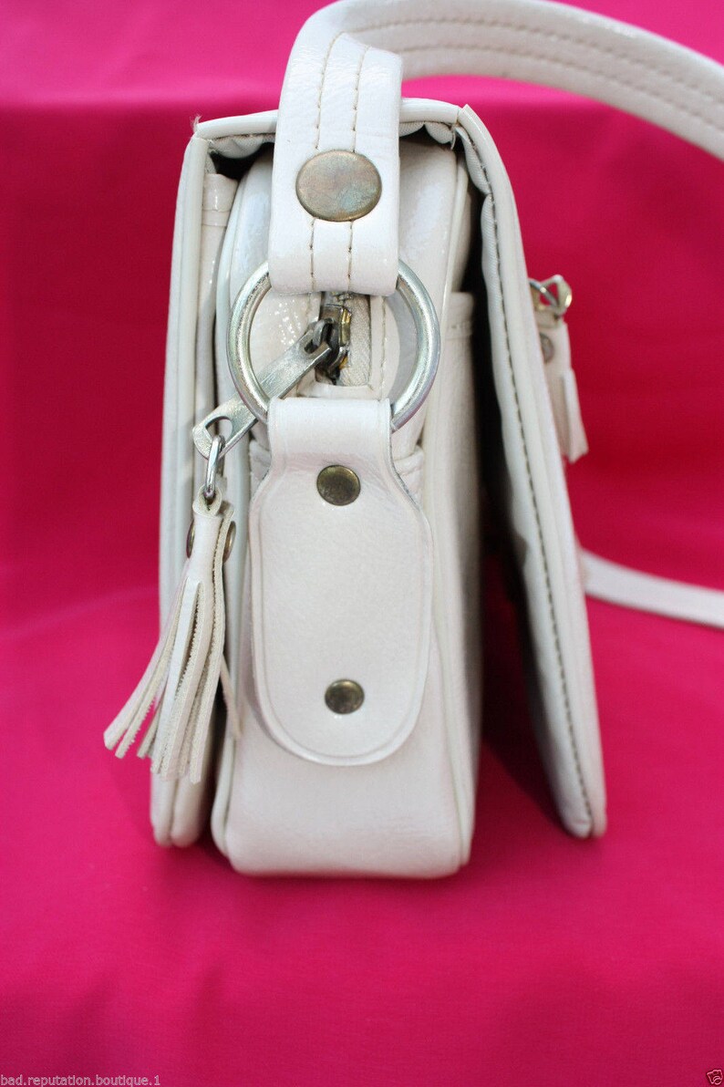 May include: A white leather shoulder bag with a zip closure, a ring detail, and a tassel. The bag has a flap with a snap closure and a small pocket on the front.