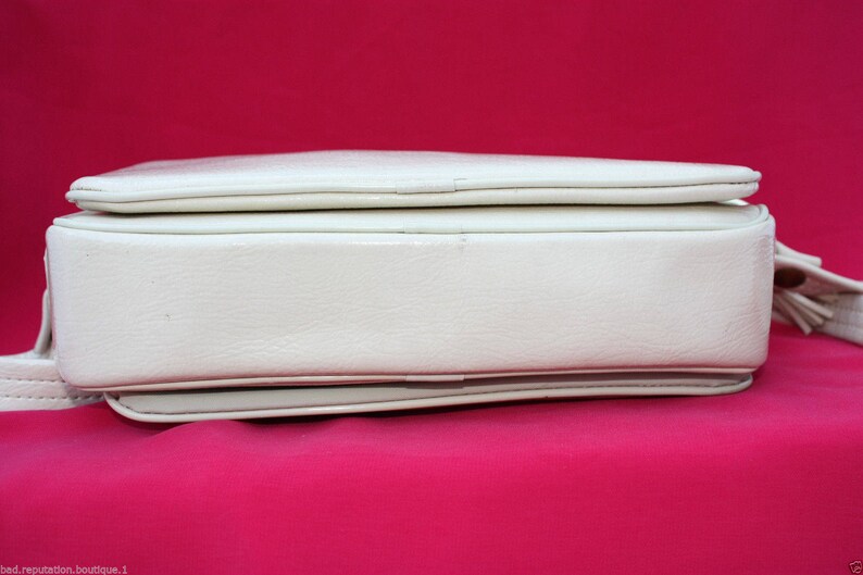 May include: A white leather handbag with a flap closure and a small tassel. The bag has a smooth, textured surface and a subtle sheen.