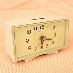 Puede incluir: Un despertador vintage de color crema con forma rectangular. La esfera es de color beige claro con números y manecillas negras. Se ven las palabras "LIGHTED DIAL". Tiene una pequeña base blanca y un cable de alimentación.