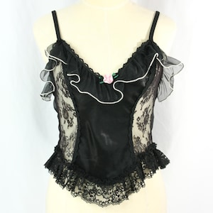 Könnte beinhalten: Schwarzes Dessous-Top mit Spitzeneinsätzen und einer Satinoberfläche. Das Top hat einen gerüschten Ausschnitt und Saum, mit zarten Perlenbesatz und einem kleinen rosa Rosenakzent. Dünne Träger vervollständigen das Design.