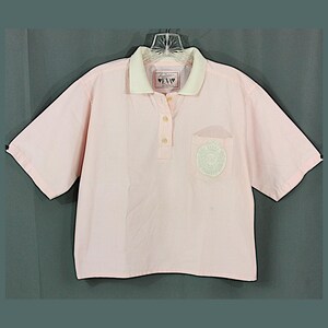 Könnte beinhalten: Hellrosa Polo-Shirt mit kurzen Ärmeln. Es hat einen weißen Kragen und eine kleine Brusttasche mit einem hellgrünen gestickten Design. Der Kragen trägt den Text "EVA".
