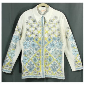 May include: White button-down shirt with a floral pattern in light blue, yellow, and gray. The shirt features a collared neckline and long sleeves. The floral design is arranged in a grid pattern across the front and sleeves.