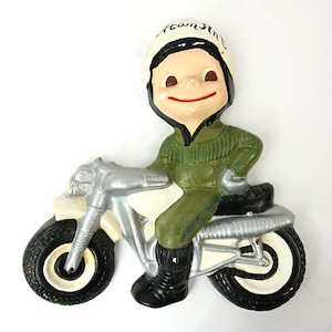 Op de afbeelding: Een keramiek beeldje van een lachende jongen die een witte helm draagt met de tekst "Atom Ant" erop, rijdend op een zilveren en zwarte motorfiets.