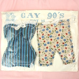 May include: A set of two fabric items in a clear plastic bag. The items are shaped like a corset and trousers. The corset is blue and white striped and the trousers are beige with a floral pattern. The bag has the text "Gay 90's" printed on it.