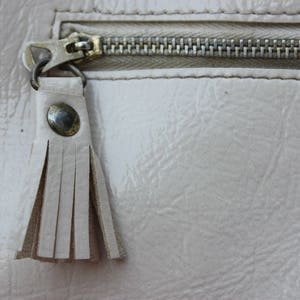 May include: A close-up of a light beige zipper with a tassel pull. The zipper is closed and the tassel is made of a soft, light beige material.