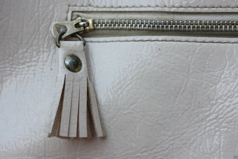 May include: A close-up of a light beige zipper with a tassel pull. The zipper is closed and the tassel is made of a soft, light beige material.