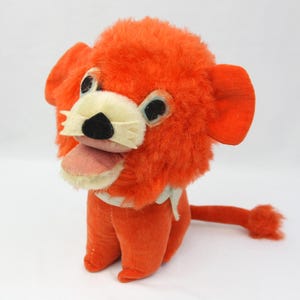 May include: Orange plush lion with a white collar, black nose, and pink tongue. The lion is sitting with its tail curled behind it.
