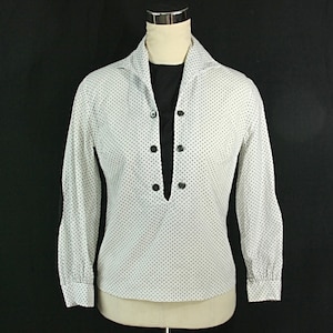 May include: A white long-sleeved blouse with black polka dots and a black mock neck collar. The blouse has a V-neckline and buttons down the front.