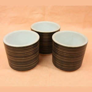May include: Three ceramic cups with a dark brown, horizontally striped exterior and a white interior. The cups are cylindrical with a slightly flared rim. The background is a solid peach color.