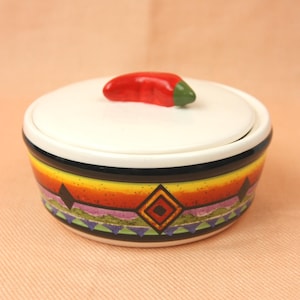 Könnte beinhalten: Eine weiße Keramikschüssel mit Deckel und einer roten Chilischote obenauf. Die Schüssel hat ein farbenfrohes südwestliches Design mit schwarzen, gelben, orangefarbenen, grünen und violetten Akzenten.