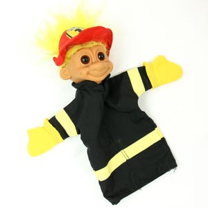 Puede incluir: Una muñeca troll vintage vestida de bombero. La muñeca tiene pelo amarillo, un gorro de bombero rojo y un uniforme negro con rayas amarillas. Los brazos de la muñeca están extendidos y tiene una expresión sonriente.