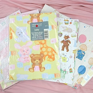 Könnte beinhalten: Ein Set mit sechs Geschenkpapierrollen zum Thema Baby. Die Rollen zeigen verschiedene Designs, darunter eine gelbe Giraffe, ein rosa Kaninchen, ein blauer Teddybär, ein blau-weiß gestreifter Teddybär, ein Baby mit einem blauen Ballon und ein Baby mit einem rosa Ballon. Die Rollen sind in durchsichtigem Plastik verpackt.