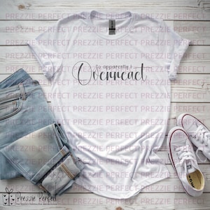 Puede incluir: Una camiseta blanca con el texto "So apparently I Overreact" en escritura negra. La camiseta se combina con vaqueros azul claro y zapatillas blancas. La imagen está sobre un fondo de madera clara.