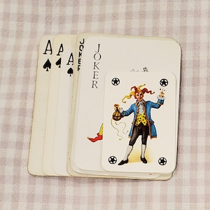 May include: A stack of playing cards featuring three aces of spades and a joker card. A smaller, separate joker card is placed on top, displaying a colorful illustration of a jester in a blue coat and yellow vest, holding a bag and small cards.