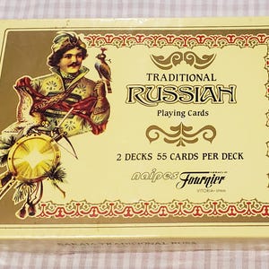 May include: A rectangular box of Traditional Russian Playing Cards by Naipes Fournier. The cream-colored box features an illustration of a person in historical attire with a bird and shield, text reading 2 DECKS 55 CARDS PER DECK, and a red border.