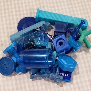 May include: A collection of various blue plastic objects, including a cylinder, a button, a small horn, and a rectangular block. The objects are in shades of blue, from light to dark, and are scattered on a checkered surface.