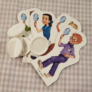 May include: A collection of game pieces featuring cartoon illustrations of children holding magnifying glasses. The game pieces are white and shaped like people. There are also several white, circular game pieces. The copyright mark "©1998 Hasbro" is visible.