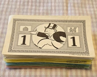 May include: A stack of Monopoly game money with a $1 bill on top. The bill features a cartoon man in a top hat running with a house and a train in the background. The text "1990 Parker Brothers" is printed at the bottom of the bill.