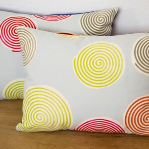 May include: Two rectangular cushions in light blue, featuring a spiral pattern in red, green, and brown. The cushions are placed on a wooden surface. The edges of the cushions have a blue trim.