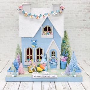 May include: A light blue Easter-themed miniature house with a white roof and decorative accents. Features a bunny figurine holding a carrot, miniature trees, and floral arrangements. The house has a garland and the text "glitteratmidnight.etsy.com".
