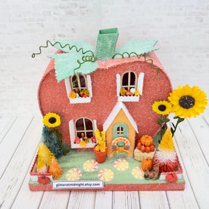 May include: A miniature pumpkin-shaped house with a glittery orange exterior, green leaves, and a stem. It features arched windows with flower boxes, a central door with a wreath, sunflowers, small trees, pumpkins, and a squirrel on a green base.