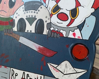May include: A wooden cutout sign featuring cartoon illustrations of horror characters in a dark blue vehicle. The design includes a white paper boat labeled S.S. Georgie, a hockey mask, a knife, and the text Be Afraid with red blood splatter.