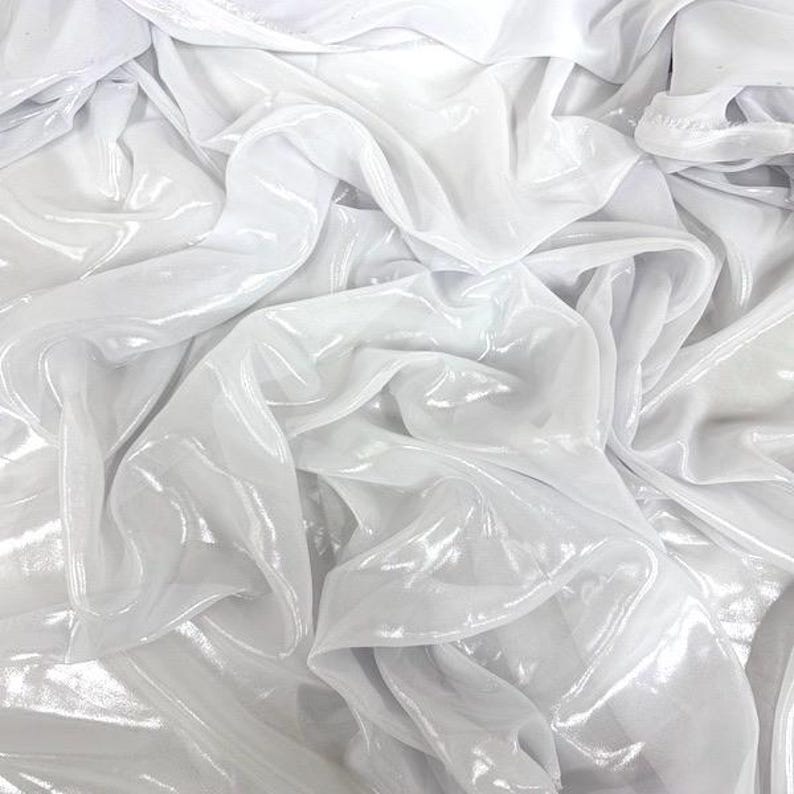 May include: Close-up of white, shimmering fabric with gentle folds and a reflective surface. The fabric is draped, creating a flowing, textured effect. The material has a subtle sheen, possibly satin or a similar finish.