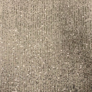 May include: A close-up of a silver fabric with a sparkly, textured surface. The fabric is made of small, round sequins that are tightly woven together.