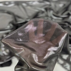 May include: Close-up of a shimmering, sheer fabric in shades of grey and silver. The fabric is draped and folded, creating a play of light and shadow. The material appears lightweight and delicate, with a subtle sheen.