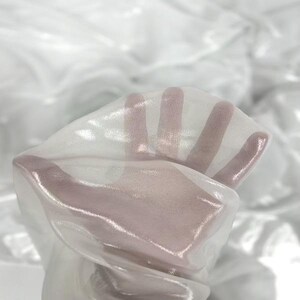 May include: Close-up of sheer white fabric, held up to the light. The fabric is translucent, allowing the hand to be visible through it. The material has a subtle sheen, creating highlights and shadows.
