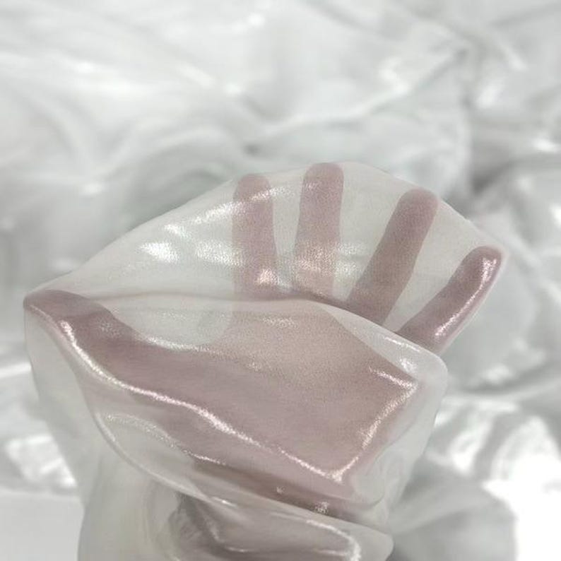 May include: Close-up of sheer white fabric, held up to the light. The fabric is translucent, allowing the hand to be visible through it. The material has a subtle sheen, creating highlights and shadows.