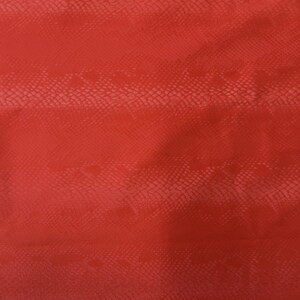 May include: A close-up of a textured, reddish-orange fabric with a snakeskin pattern. The material has a subtle sheen and a repeating scale-like design, creating a reptile skin effect. The color is a warm, earthy tone.