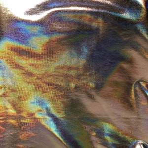 May include: Close-up of iridescent fabric with a metallic sheen. The fabric is wrinkled and has a textured surface. The colors are a mix of blue, green, and gold.