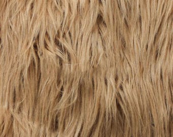 May include: A close-up view of long, shaggy faux fur fabric in a light tan or beige color. The texture consists of dense, flowing synthetic fibers that create a layered, multi-directional appearance with subtle variations in golden brown tones.