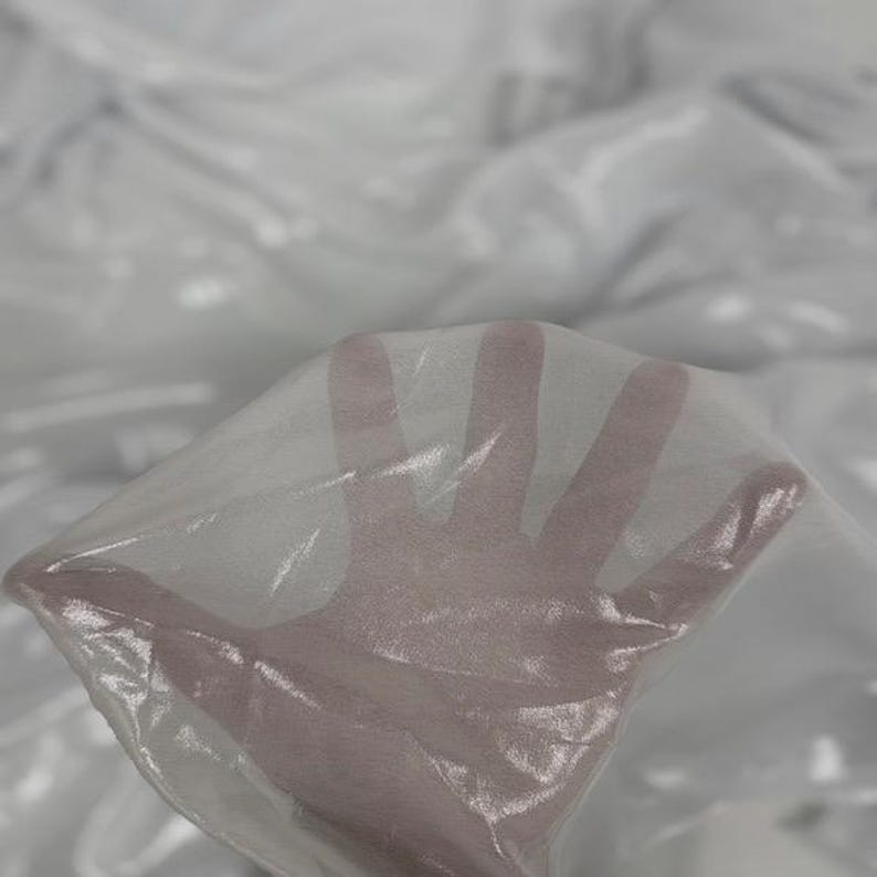 May include: Close-up of a sheer white fabric, possibly organza or chiffon, with a subtle sheen. The fabric is draped and slightly translucent, with a hand visible through the material. The overall effect is delicate and airy.