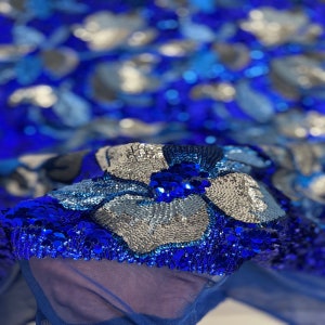 May include: Close-up of a blue and silver sequined fabric with a floral design. The sequins are arranged in a pattern of flowers and leaves.