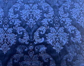 May include: A deep blue velvet fabric featuring a repeating damask pattern. The design consists of intricate, symmetrical floral and scrollwork motifs that are slightly raised, creating a textured appearance against the smooth base.
