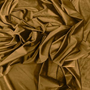 May include: A length of solid brown velvet fabric featuring a soft, dense pile and a subtle sheen. The material is draped in deep, flowing folds that create varying light and shadow across the smooth, textured surface of the textile.