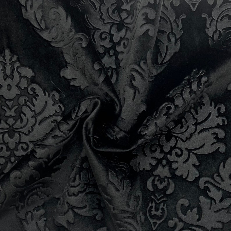 Pode incluir: Close-up de tecido preto com um padr&atilde;o de damasco em relevo. O tecido est&aacute; dobrado, criando sombras e destacando a textura do design. O padr&atilde;o apresenta folhas e motivos florais.
