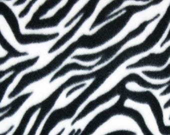 May include: A soft fabric featuring a high-contrast zebra print pattern with bold, irregular black stripes set against a clean white background. The surface texture appears plush and fuzzy, displaying a repeating animal-inspired motif.