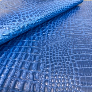 May include: Close-up of a blue crocodile embossed leather fabric. The fabric has a textured, scaly pattern.
