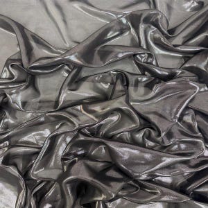 May include: A close-up of a metallic grey fabric with a shimmering finish. The material is draped, creating soft folds and highlights. The fabric's texture is fluid and reflects light, giving it a luxurious appearance. The overall effect is one of elegance and movement.