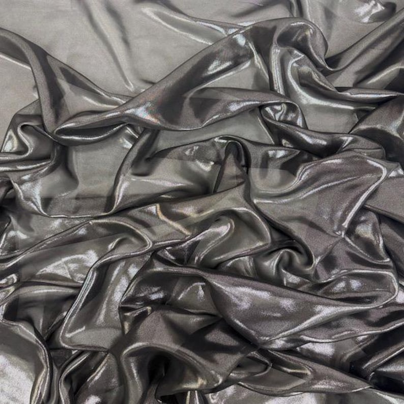 May include: A close-up of a metallic grey fabric with a shimmering finish. The material is draped, creating soft folds and highlights. The fabric's texture is fluid and reflects light, giving it a luxurious appearance. The overall effect is one of elegance and movement.