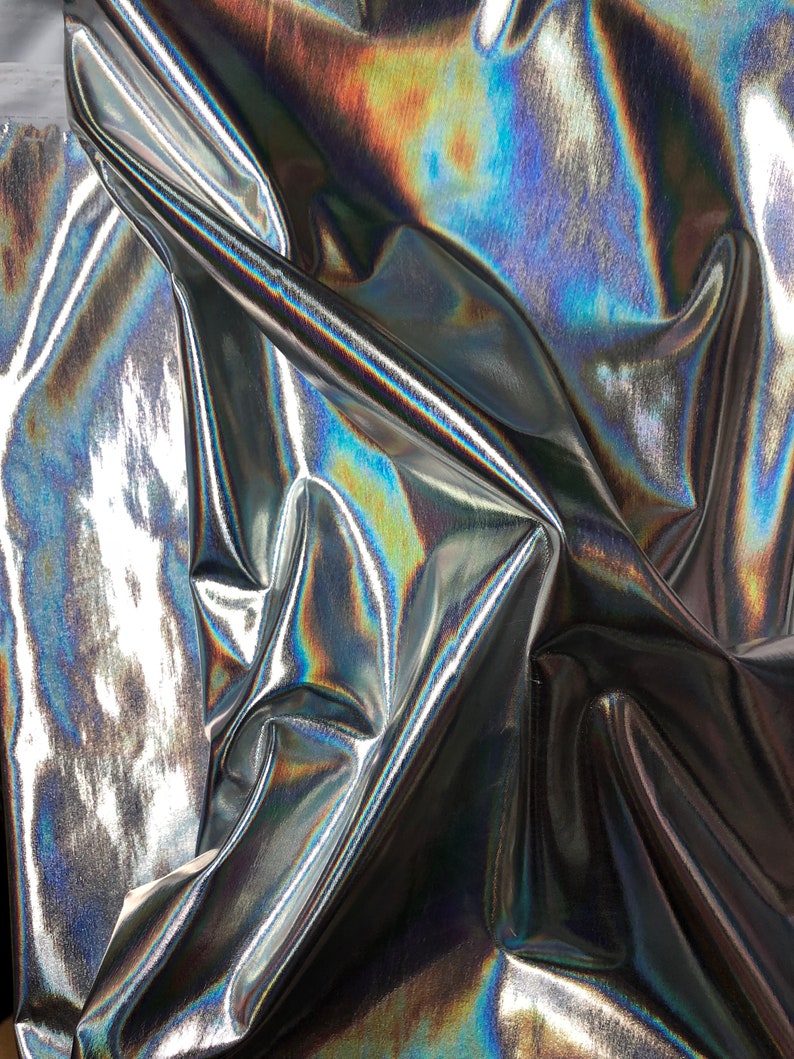 May include: Close-up of iridescent silver fabric with a textured surface. The fabric is folded and draped, creating a dynamic and abstract pattern.