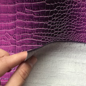 May include: Close-up of a purple faux leather material with a crocodile skin texture. The material is being lifted to reveal a white, felt-like backing. The surface has a glossy finish and a detailed pattern.
