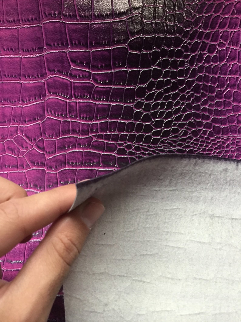 May include: Close-up of a purple faux leather material with a crocodile skin texture. The material is being lifted to reveal a white, felt-like backing. The surface has a glossy finish and a detailed pattern.