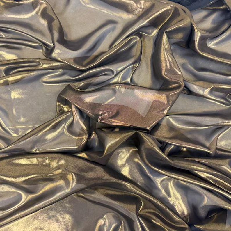 May include: Close-up of shimmering fabric with a metallic sheen. The fabric is a blend of silver and gold, with soft folds and highlights creating a luxurious appearance. The material appears lightweight and flowing, ideal for fashion or decor.