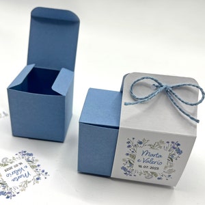 Può includere: Due scatole portaconfetti blu con coperchi bianchi. Una scatola è aperta e l'altra è chiusa con un fiocco di spago blu e bianco. La scatola chiusa presenta un disegno floreale con il testo "Marta e Valerio 16.07.2023".