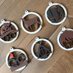 Può includere: Ornamenti natalizi in legno con cornici circolari bianche. Gli ornamenti presentano ritagli marroni e neri di un cappello parlante, un cervo, il numero 9 e 3/4, capelli e occhiali, un fulmine e il simbolo dei Doni della Morte.