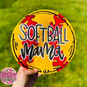 May include: A yellow softball-shaped decoration with the words "SOFTBALL Mama" in white and black. Red stitching-like accents are visible. The decoration is held against a blurred green background.